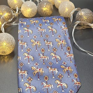Può includere: Una cravatta blu con un motivo ripetuto di cavalli da giostra nei colori crema, rosso e oro. I cavalli sono raffigurati in varie posizioni, ciascuno attaccato a un palo verticale sormontato da un elemento floreale decorativo.