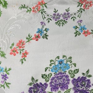 May include: White fabric with an embroidered floral pattern. The design features clusters of purple, pink, and blue flowers with green leaves. A subtle, embossed pattern is visible in the background.