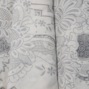 Pode incluir: Close-up de um têxtil branco com intrincados bordados florais cinzentos. O design apresenta grandes flores, folhas e padrões geométricos, criando um visual detalhado e elegante. O tecido parece ser linho ou um material semelhante.