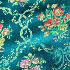 May include: Close-up of teal fabric with a floral pattern. The design features bouquets of red, pink, and yellow flowers, along with green bows and vines. The fabric has a textured appearance, suggesting a brocade or damask weave.