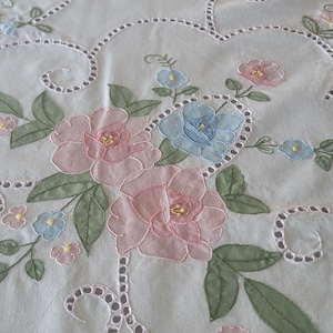 Peut inclure: Une nappe en tissu blanc ornée de broderies ajourées complexes le long de bordures incurvées. Le textile est décoré de motifs floraux dans des tons pastel doux de rose, bleu clair et vert sauge, avec des points détaillés sur les pétales et feuilles.