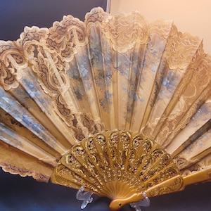 Op de afbeelding: Een vouwwaaier met een lichtbruine, uitvoerig gesneden houten beschermkap en baleinen. Het waaierblad is gemaakt van beige kant met een geschulpte rand en een centraal paneel met een zacht, gedempt blauw en bruin bloemmotief.