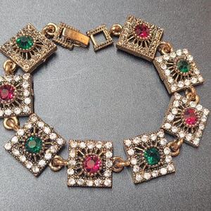 Peut inclure: Un bracelet à maillons en métal doré composé de huit maillons carrés. Chaque maillon est bordé de petits strass transparents et contient une pierre précieuse ronde centrale, alternant entre les couleurs rouge et verte. Fermoir en métal.