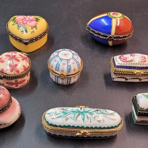 Könnte beinhalten: Eine Sammlung von acht kleinen Schmuckdosen aus Porzellan in verschiedenen Formen und Designs, darunter ein gelbes Herz, ein blau-rotes Ei sowie mehrere rechteckige und runde Behälter mit Blumenmustern und goldfarbenen Metallscharnieren.