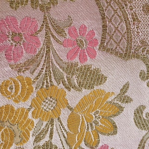 May include: Close-up of a floral fabric with a repeating pattern. The design features pink and yellow flowers with green leaves and stems on a light pink background. The fabric has a textured appearance.