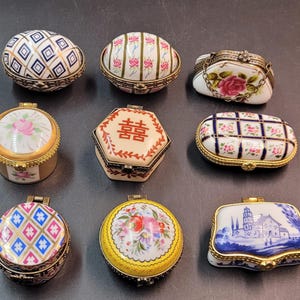 Pode incluir: Uma coleção de nove pequenas caixas de porcelana para bugigangas com fechos de metal articulados. As caixas apresentam várias formas, incluindo ovais, redondas, hexagonais e retangulares, decoradas com padrões florais, grades geométricas e um símbolo chinês de felicidade dupla.
