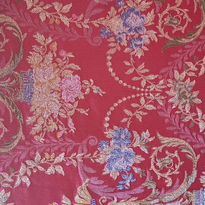 May include: Red fabric with an ornate floral pattern. The design features gold, pink, and blue flowers and foliage on a red background. The fabric has a textured appearance, suggesting a brocade or damask material.
