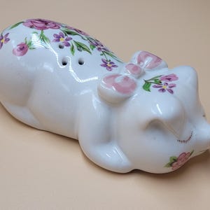 Könnte beinhalten: Ein weißer Salz- und Pfefferstreuer in Schweineform aus Keramik. Das Schwein ist mit rosa und lila Blumenmustern verziert und hat zwei Löcher auf dem Rücken. Das Schwein hat eine rosa Schleife auf dem Kopf und geschlossene Augen.