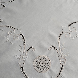 Pode incluir: Uma toalha de mesa branca com bordados intrincados e medalhões circulares de crochê. O design inclui motivos florais em espiral e padrões vazados que criam uma superfície delicada e texturizada sobre o tecido da toalha de mesa.