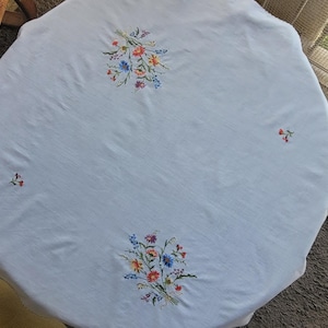 Puede incluir: Un mantel redondo de color blanco que presenta dos ramos de flores bordados en tonos rojo, azul, amarillo y verde, situados en la parte superior e inferior, con dos pequeñas ramitas florales bordadas en los lados izquierdo y derecho.