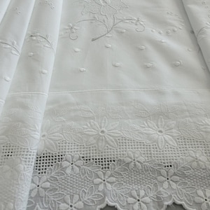 Peut inclure: Un textile en tissu blanc présentant une broderie florale complexe et un travail ajouré le long de la bordure festonnée. La surface affiche un motif répétitif de petits points en relief et de délicats brins floraux brodés.