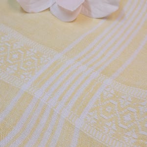 Op de afbeelding: Een lichtgeel textiel met witte verticale strepen en een decoratief geometrisch geweven randpatroon. Een grote, lichtroze kunstmatige magnolia-bloem met gelaagde bloemblaadjes rust op het oppervlak van de gevouwen stof.