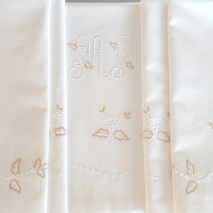Può includere: Un runner in tessuto bianco con una lettera monogramma M ricamata al centro in filo bianco. Il runner è decorato con motivi ricamati di foglie e viticci in marrone chiaro lungo i bordi e motivi floreali sparsi sulla superficie.