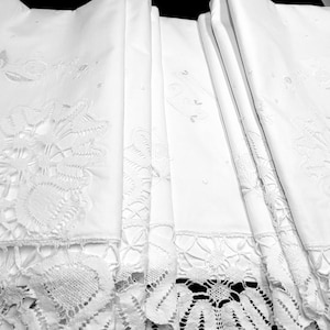 Puede incluir: Un conjunto de mantelería blanca rectangular con intrincados bordados calados y anchos bordes de encaje festoneado. La tela muestra delicados motivos florales cosidos en hilo blanco, con patrones de calados a lo largo de los bordes de cada pieza.