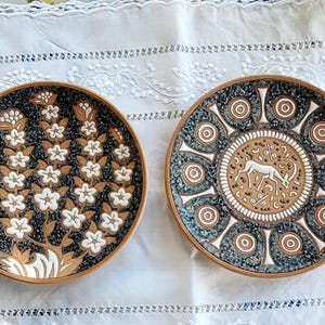 May include: Two round ceramic plates with terracotta rims. The left plate features a black background with white floral branches. The right plate displays a central white gazelle surrounded by concentric circles and intricate black and white patterns.