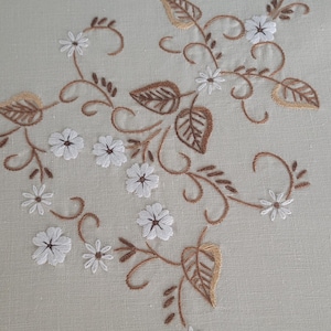May include: A light beige fabric featuring delicate embroidery of white flowers with dark centres and brown swirling vines with leaves. The floral pattern is arranged in a flowing, organic design across the surface of the textile material.