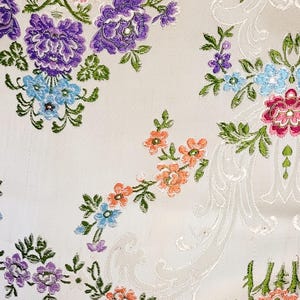 May include: Close-up of a white fabric with an embroidered floral pattern. The flowers are in shades of red, purple, blue, and orange, with green leaves. The background has a subtle, textured design.