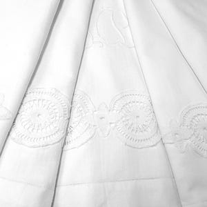 Può includere: Tessuto bianco con ricami in pizzo circolare e intricato lungo i bordi. Il tessuto è piegato, creando un effetto a strati. Il design in pizzo presenta motivi floreali e geometrici.