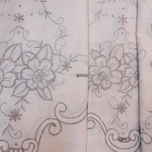 Könnte beinhalten: Ein weißer Stoff mit aufwendiger hellbrauner Blumenstickerei. Das Design zeigt detaillierte Blüten, Blätter und geschwungene Rankenmuster, die in einem sich wiederholenden Motiv über die Oberfläche angeordnet sind, ergänzt durch dekorative Bogenkanten.