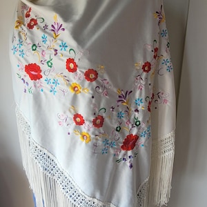Puede incluir: Un chal color blanco roto con bordados florales multicolores en tonos rojo, amarillo, azul y morado. La prenda está rematada con un borde ancho de encaje de ganchillo intrincado y largos flecos fluidos en el borde inferior.