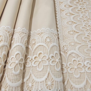Pode incluir: Close-up de tecido creme dobrado com intrincada borda de renda branca. A renda apresenta um design floral e bordas onduladas. O tecido parece ser uma toalha de mesa ou têxtil semelhante.