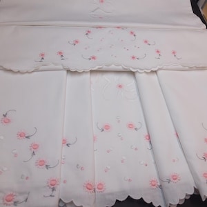 Puede incluir: Un conjunto de tela blanca con delicados bordados florales en rosa con tallos y hojas verdes. Las piezas tienen bordes festoneados y están decoradas con pequeños puntos blancos dispersos por la superficie, creando un conjunto textil detallado y coherente.
