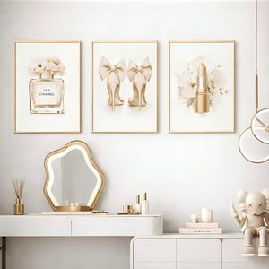 Op de afbeelding: Drie ingelijste wandkunstwerken met gouden frames. De kunst toont een parfumfles met bloemen, hoge hakken met strikken en een lippenstift met bloemen. Het kleurenpalet is neutraal met gouden accenten.