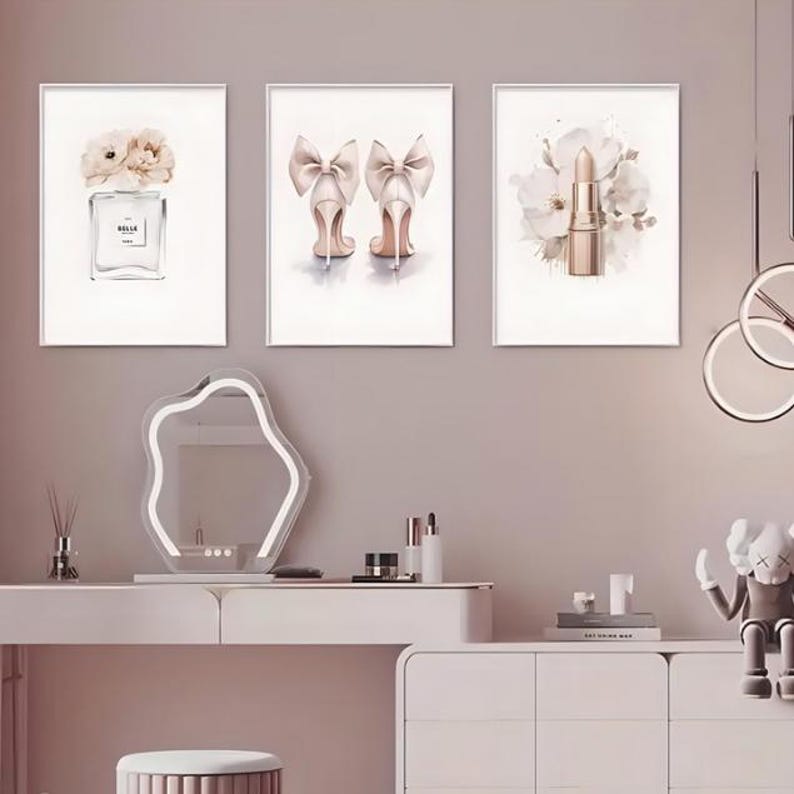 May include: Three framed watercolor prints. One features a perfume bottle with flowers, another shows high heels with bows, and the third depicts a lipstick with floral accents. The prints are displayed above a vanity.
