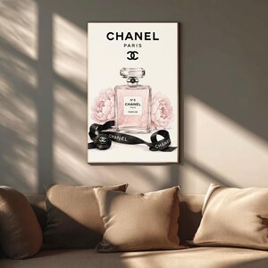 May include: A rectangular wall art print featuring a clear glass perfume bottle labeled Chanel N&deg;5 Parfum, centered above two soft pink peony flowers. A black ribbon with the Chanel logo and text is draped in front, set against a cream background.