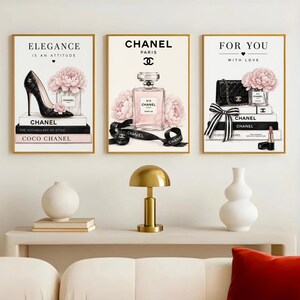 May include: A set of three wall art prints featuring fashion-themed illustrations. The left print shows a black high heel, pink flowers, and books. The center print displays a perfume bottle and flowers. The right print depicts a handbag, flowers, and lipstick.