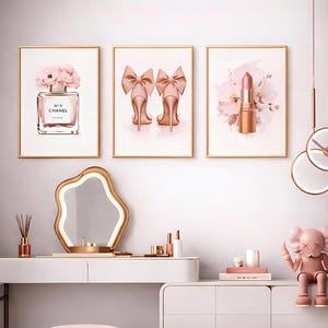 Könnte beinhalten: Drei gerahmte Kunstdrucke mit goldenen Rahmen. Die Drucke zeigen einen Parfümflakon, High Heels mit Schleifen und einen Lippenstift, alles in Rosatönen. Die Kunst wird über einem weißen Schminktisch mit Spiegel und dekorativen Gegenständen ausgestellt.