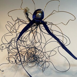 May include: A wire sculpture of a figure with a blue ribbon and a white flower. The figure is made of gold and black wire and is standing on a white surface.