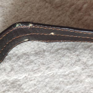 Peut inclure: Une lani&egrave;re en cuir marron fonc&eacute; avec des coutures orange visibles sur les bords. La lani&egrave;re pr&eacute;sente des signes d'usure, notamment de petites zones o&ugrave; la finition fonc&eacute;e s'est effac&eacute;e pour r&eacute;v&eacute;ler un mat&eacute;riau plus clair sur la section courb&eacute;e.