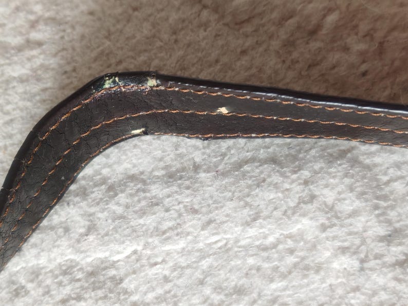 Peut inclure: Une lani&egrave;re en cuir marron fonc&eacute; avec des coutures orange visibles sur les bords. La lani&egrave;re pr&eacute;sente des signes d'usure, notamment de petites zones o&ugrave; la finition fonc&eacute;e s'est effac&eacute;e pour r&eacute;v&eacute;ler un mat&eacute;riau plus clair sur la section courb&eacute;e.