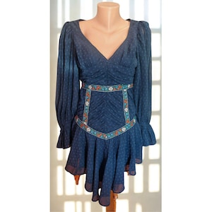 May include: A navy blue dress with a V-neck and long sleeves. The dress has a floral embroidered belt and a ruffled skirt. The fabric has a subtle polka dot pattern. The dress is displayed on a mannequin.