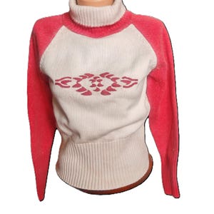 Könnte beinhalten: Ein cremefarbener Rollkragenpullover mit korallenfarbenen Raglanärmeln und einem passenden geometrischen Design auf der Vorderseite. Der Pullover hat einen gerippten Saum und Bündchen und wird auf einer Schaufensterpuppe präsentiert.