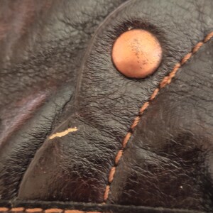 Peut inclure: Un gros plan sur du cuir marron fonc&eacute; pr&eacute;sentant une surface textur&eacute;e avec un grain visible. Un seul rivet m&eacute;tallique rond de couleur cuivre est fix&eacute; pr&egrave;s d'une couture r&eacute;alis&eacute;e avec un fil beige &eacute;pais et contrast&eacute;. Une usure est visible.