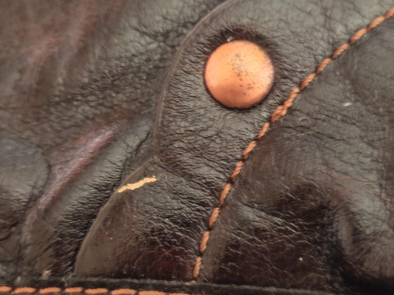 Peut inclure: Un gros plan sur du cuir marron fonc&eacute; pr&eacute;sentant une surface textur&eacute;e avec un grain visible. Un seul rivet m&eacute;tallique rond de couleur cuivre est fix&eacute; pr&egrave;s d'une couture r&eacute;alis&eacute;e avec un fil beige &eacute;pais et contrast&eacute;. Une usure est visible.