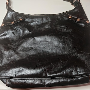 Peut inclure: Un sac &agrave; bandouli&egrave;re noir de style hobo avec une finition brillante et textur&eacute;e. Le sac pr&eacute;sente des empi&egrave;cements en cuir festonn&eacute;s sur les coins sup&eacute;rieurs, chacun fix&eacute; par trois rivets m&eacute;talliques cuivr&eacute;s. Il poss&egrave;de une sangle unique.