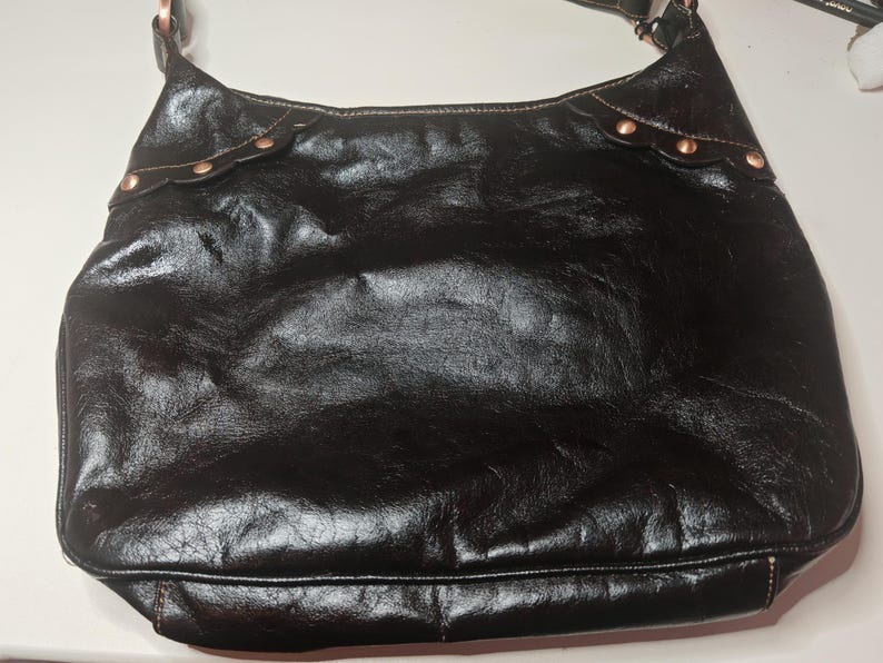 Peut inclure: Un sac &agrave; bandouli&egrave;re noir de style hobo avec une finition brillante et textur&eacute;e. Le sac pr&eacute;sente des empi&egrave;cements en cuir festonn&eacute;s sur les coins sup&eacute;rieurs, chacun fix&eacute; par trois rivets m&eacute;talliques cuivr&eacute;s. Il poss&egrave;de une sangle unique.