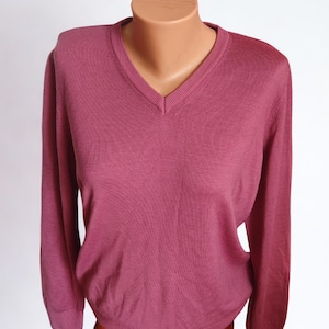 Può includere: Un maglione color malva con scollo a V e maniche lunghe. La maglia è esposta su un manichino. Il maglione ha un design classico e una trama a coste. Il colore è un viola-rossastro smorzato.