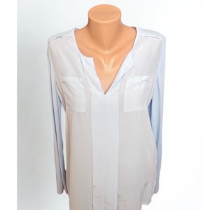 May include: A pale blue long-sleeved blouse with a V-neck and two front pockets. The blouse features a central vertical panel and is displayed on a mannequin. The material appears to be lightweight.