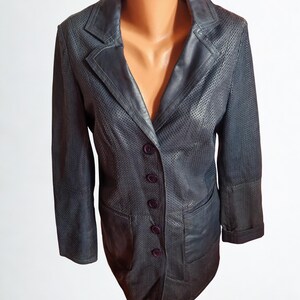 Peut inclure: Un blazer bleu marine foncé avec un design perforé. Le blazer a un revers cranté, une fermeture à trois boutons et deux poches avant. Les manches sont retroussées et le blazer est présenté sur un mannequin.