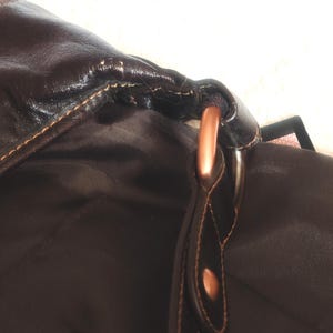 Peut inclure: Un sac marron fonc&eacute; avec un ext&eacute;rieur lisse et des coutures apparentes, reli&eacute; &agrave; une sangle par un anneau m&eacute;tallique de couleur cuivre. L'int&eacute;rieur est doubl&eacute; d'un tissu marron fonc&eacute; et un petit rivet cuivr&eacute; est visible sur la fixation de la sangle.