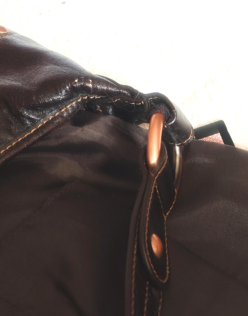 Peut inclure: Un sac marron fonc&eacute; avec un ext&eacute;rieur lisse et des coutures apparentes, reli&eacute; &agrave; une sangle par un anneau m&eacute;tallique de couleur cuivre. L'int&eacute;rieur est doubl&eacute; d'un tissu marron fonc&eacute; et un petit rivet cuivr&eacute; est visible sur la fixation de la sangle.