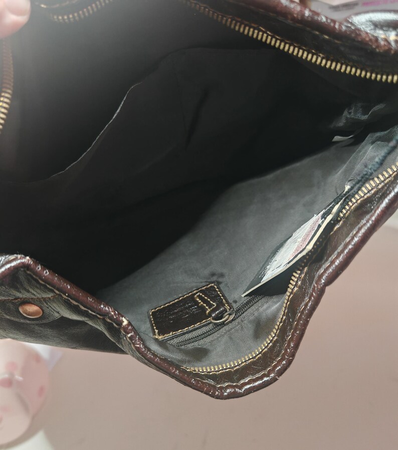 Peut inclure: Un sac &agrave; main en cuir marron fonc&eacute; avec une fermeture &eacute;clair sur le dessus et des d&eacute;tails dor&eacute;s. L'int&eacute;rieur est doubl&eacute; de tissu noir et comprend une poche zipp&eacute;e avec une &eacute;tiquette en cuir ainsi qu'une poche ouverte pour le rangement.