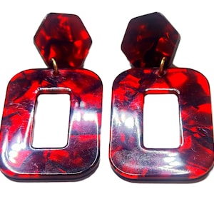 Può includere: Un paio di orecchini in acrilico marmorizzato rosso e nero. Ogni orecchino ha un perno esagonale e una goccia rettangolare con un centro ritagliato. Gli orecchini sono collegati con un elemento metallico color oro.