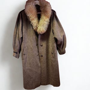 Peut inclure: Un manteau en tweed marron avec un grand col en fourrure duveteuse brun clair. Le manteau a une fermeture à boutons, deux poches et des manches longues. Le manteau est suspendu à un cintre blanc sur fond blanc.
