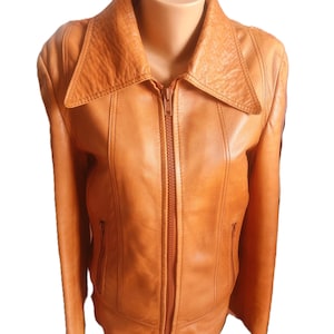 May include: A tan leather jacket with a wide collar and a full-length zip. The jacket has two zipped pockets and vertical seams on the front. The jacket is displayed on a mannequin.