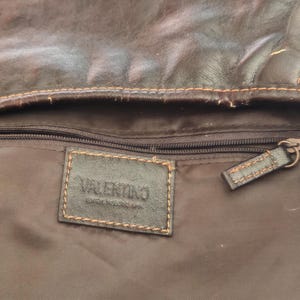 Peut inclure: L'int&eacute;rieur d'un sac en cuir marron fonc&eacute; avec une &eacute;tiquette rectangulaire en cuir portant l'inscription VALENTINO en relief. La doublure est en tissu marron fonc&eacute; avec une poche zipp&eacute;e horizontale, une tirette en cuir et des coutures orange.